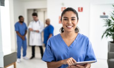Medical professional smiling to the camera with other medical professionals in the background, all wearing scrubs