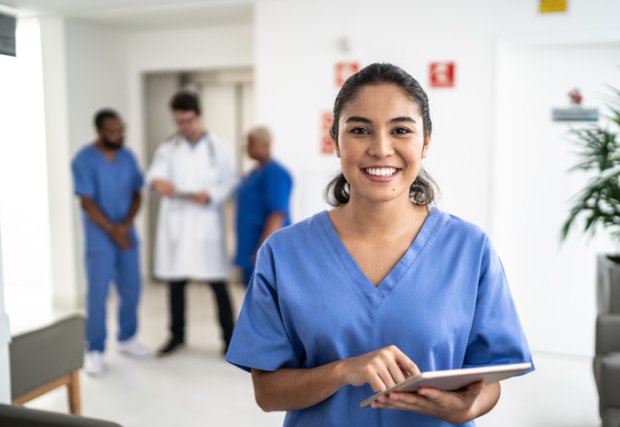 Profesional médico sonriendo a la cámara con otros profesionales médicos en el fondo, todos con batas