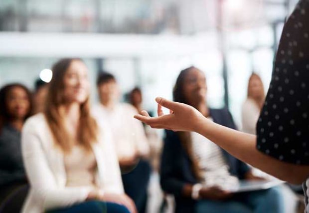 Stock image of a blurred out crowd at a lecture with the focus on the speaker's hand