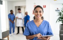 Profesional médico sonriendo a la cámara con otros profesionales médicos en el fondo, todos con batas