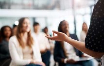 Stock image of a blurred out crowd at a lecture with the focus on the speaker's hand